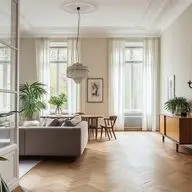 Elegant living room featuring herringbone parquet flooring, high ceilings with ornate moldings, a crystal chandelier, tall windows with sheer curtains, a grey sofa, mid-century modern furniture, and tropical plants adding natural elements to the space