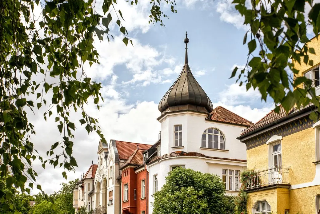 Historic European buildings with ornate roofs and colorful facades under a partly cloudy sky, framed by leafy branches.