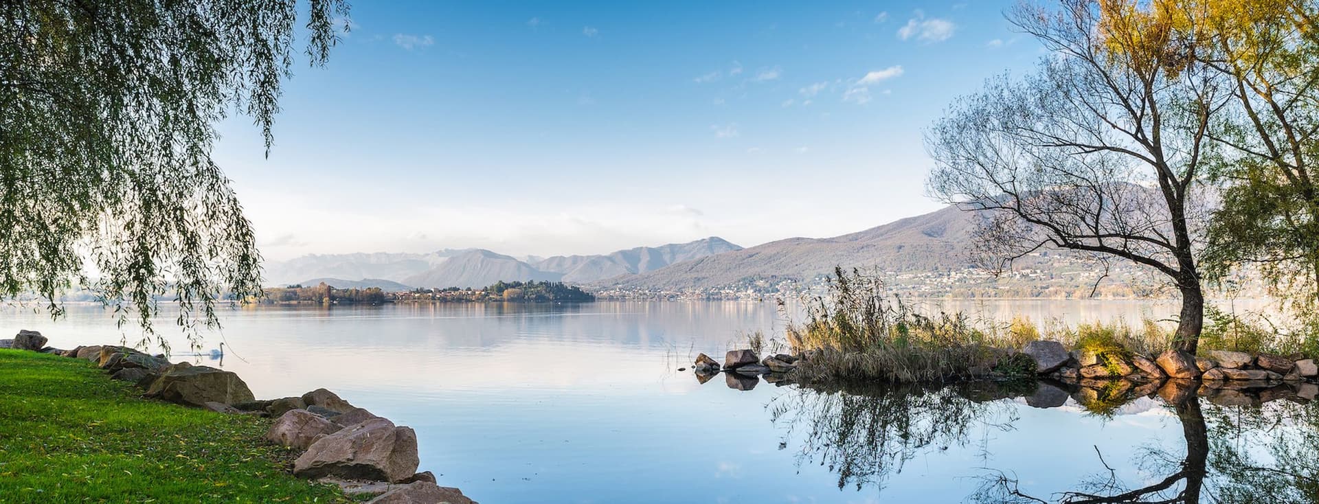 Eine friedvolle Seelandschaft Eine vollkommen ruhige Seeszene mit spiegelglatter Wasserfläche, die den tiefblauen Himmel und vereinzelte Wolken perfekt reflektiert. Im Vordergrund erstreckt sich ein grasiger Uferstreifen, gesäumt von Felsen. Bäume mit herbstlichem Laub rahmen das Bild: Eine Trauerweide auf der linken Seite und ein Baum mit goldenen Blättern auf der rechten Seite. Der spiegelglatte See erstreckt sich bis zu fernen Bergen, wobei eine kleine Insel im mittleren Hintergrund sichtbar ist. Das ruhige Wasser erzeugt perfekte Spiegelbilder der umliegenden Landschaft und schafft eine symmetrische Komposition, getaucht in weiches Morgen- oder Abendlicht.