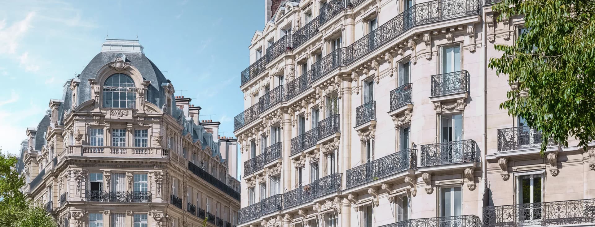 Elegant Parisian buildings with ornate balconies under a clear blue sky, surrounded by lush green trees.