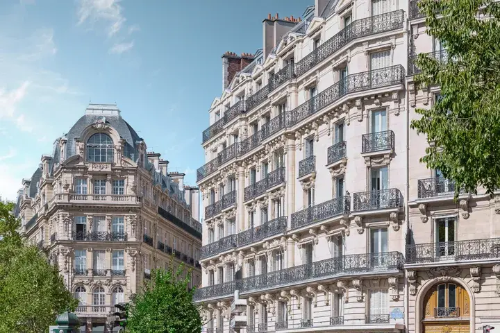 Elegant Parisian buildings with ornate balconies under a clear blue sky, surrounded by lush green trees.