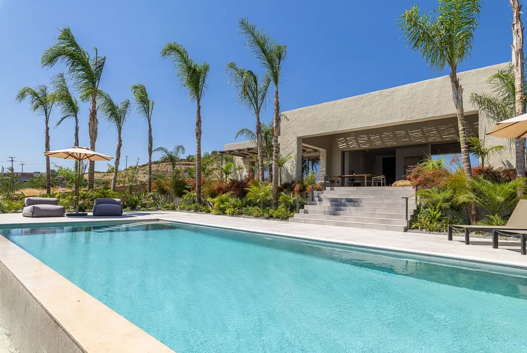 Modern beige villa with a rectangular pool, tall palm trees, and an open shaded patio with seating.