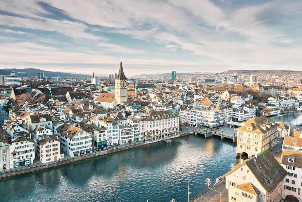 Aerial view of Zurich, Switzerland, showcasing historic buildings, a prominent church tower, and the Limmat River under a partly cloudy sky.