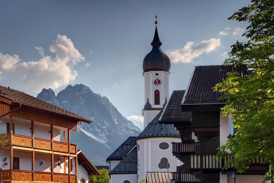 Scenic view of a church with a tall steeple against a backdrop of mountains and blue sky, surrounded by wooden buildings and lush greenery.