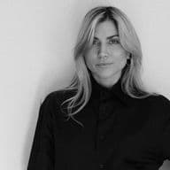 Ασπρόμαυρο πορτρέτο μιας ξανθιάς γυναίκας με μαύρο πουκάμισο με κουμπιά μπροστά σε έναν λευκό τοίχο.