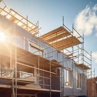 Sunlit construction site with scaffolding on a building, clear blue sky, and a few clouds in the background.