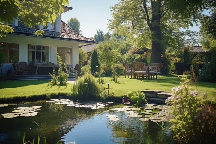 Lush garden witÜppiger Garten mit Teich, umgeben von Bäumen und Sträuchern, angrenzend an ein Haus mit Innenhof. Die Gartenmöbel stehen unter einem großen Baum.h a pond, surrounded by trees and shrubs, adjacent to a house with a patio. Outdoor furniture is set up under a large tree.