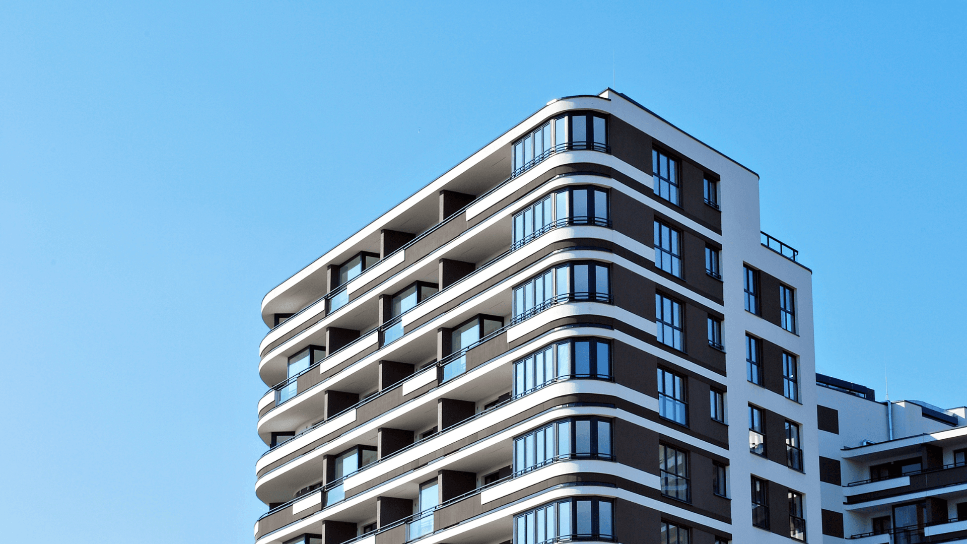Edifici d'apartaments modern amb balcons corbats amb un cel blau clar de fons. L'edifici és marró i blanc.