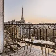 Balcony view of the Eiffel Tower at sunset, with a lounge chair and table holding a book and plant, framed by open French doors.