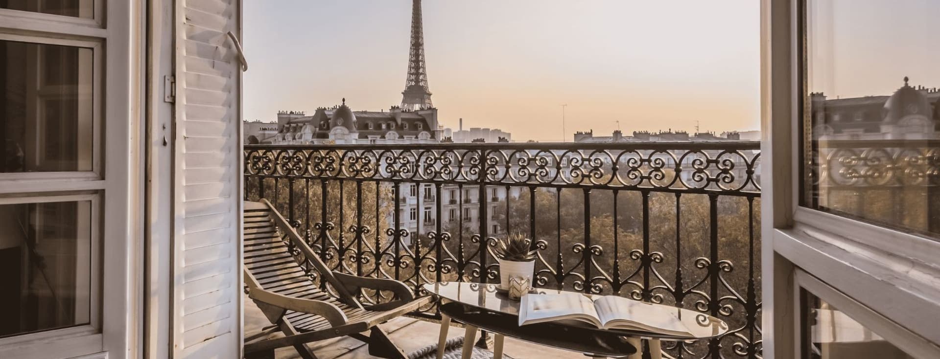 Balcony view of the Eiffel Tower at sunset, with a lounge chair and table holding a book and plant, framed by open French doors.