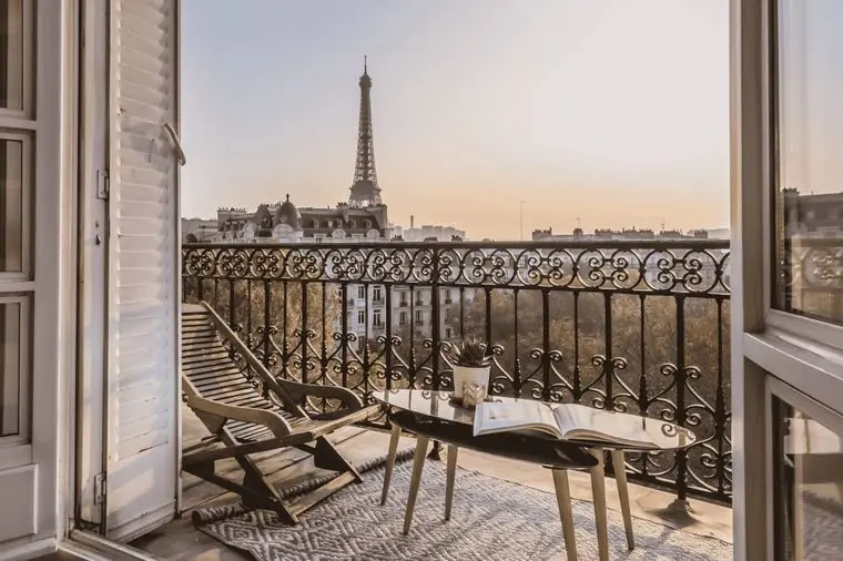 Balcony view of the Eiffel Tower at sunset, with a lounge chair and table holding a book and plant, framed by open French doors.