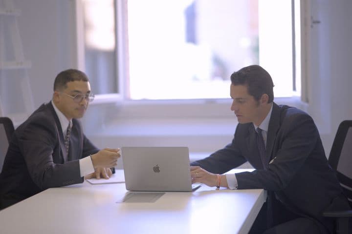 Dois homens de terno sentam-se a uma mesa branca, um usando um laptop, o outro apontando para papéis. Uma janela está no fundo.