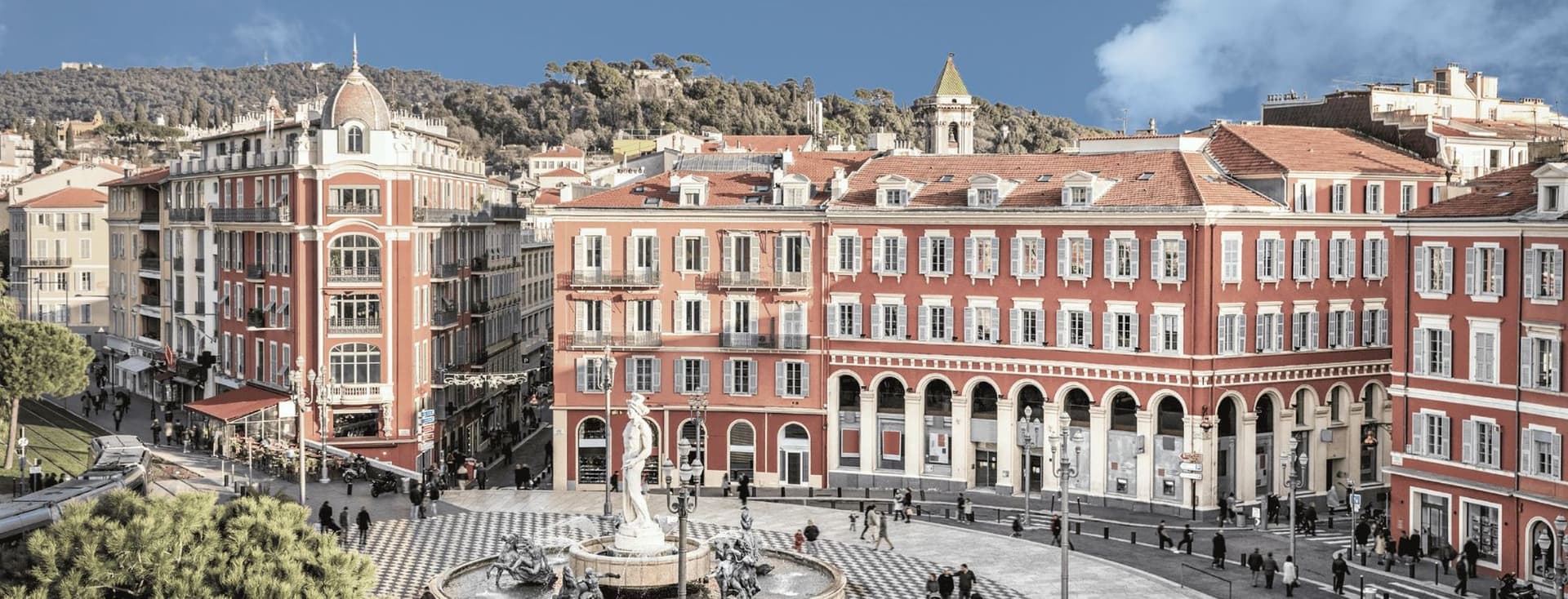 A scenic view of a European plaza with a central fountain, surrounded by red buildings with arched windows and a patterned circular pavement.