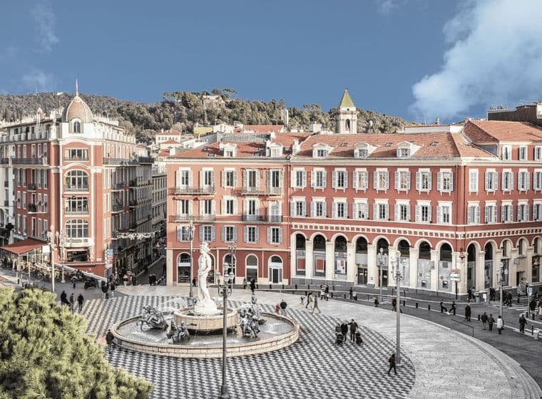 A scenic view of a European plaza with a central fountain, surrounded by red buildings with arched windows and a patterned circular pavement.