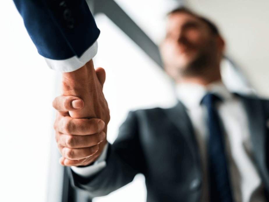 Nahaufnahme von zwei Personen, die sich in einem professionellen Umfeld die Hand geben; eine Hand ist scharf fokussiert, während die andere im Hintergrund unscharf erscheint. KI-generiertes Bild