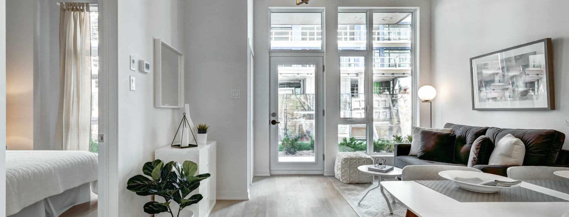 Luminoso y moderno interior de apartamento con paredes blancas, pisos de madera clara y grandes ventanas. Un sofá gris y una mesa de comedor blanca amueblan el espacio.