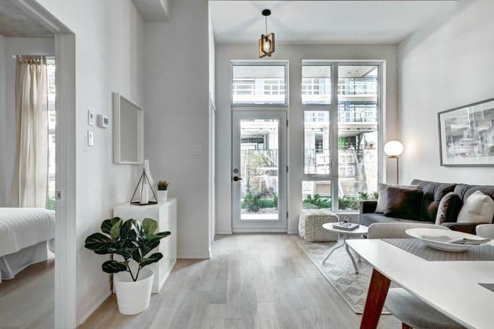 Luminoso y moderno interior de apartamento con paredes blancas, pisos de madera clara y grandes ventanas. Un sofá gris y una mesa de comedor blanca amueblan el espacio.