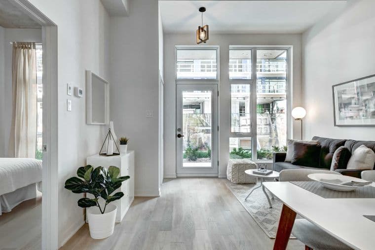 Luminoso y moderno interior de apartamento con paredes blancas, pisos de madera clara y grandes ventanas. Un sofá gris y una mesa de comedor blanca amueblan el espacio.