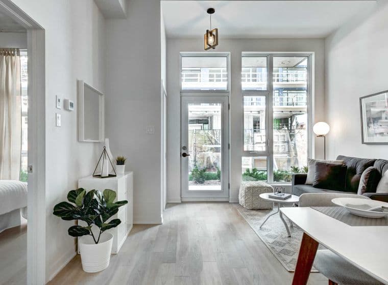 Luminoso y moderno interior de apartamento con paredes blancas, pisos de madera clara y grandes ventanas. Un sofá gris y una mesa de comedor blanca amueblan el espacio.