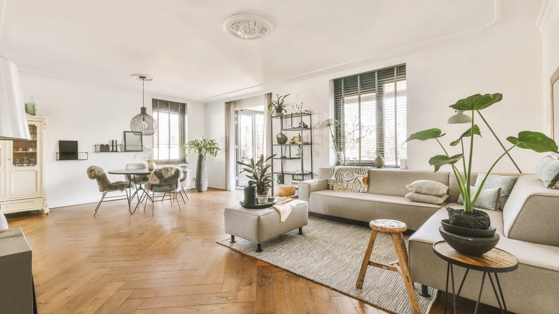 Spacious Dubai apartment living room with light wood floors, gray sofas, plants, and a dining area with a round table and chairs, under ample natural light.