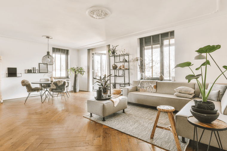 Spacious Dubai apartment living room with light wood floors, gray sofas, plants, and a dining area with a round table and chairs, under ample natural light.
