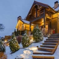 Una casa de madera de dos pisos con luces en una colina nevada al atardecer. Unos escalones de piedra conducen a la casa.