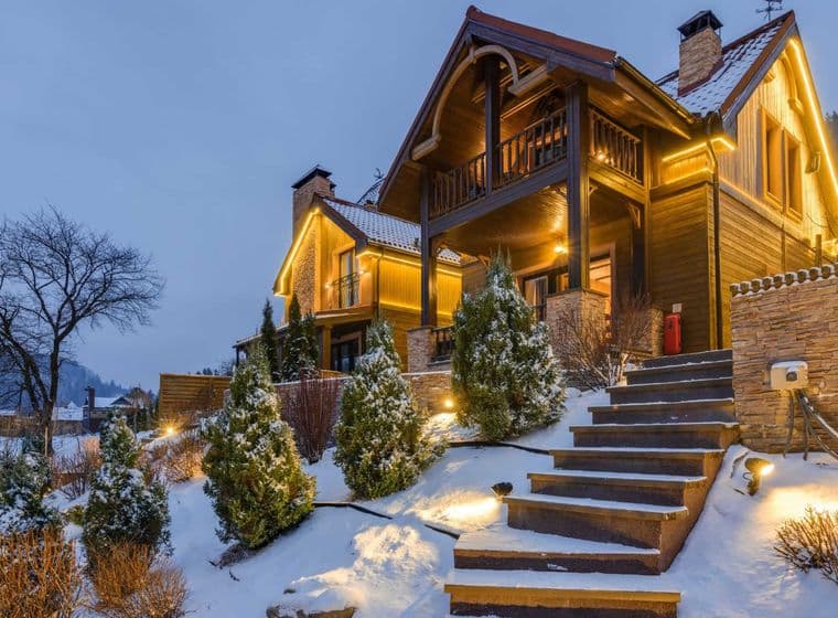 A two-story wooden house with lights on a snowy hill at dusk. Stone steps lead up to the house.