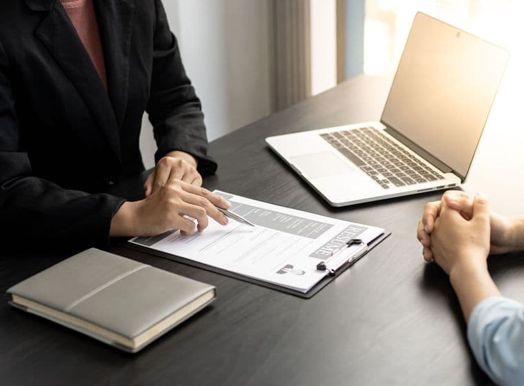 Una persona amb una americana negra revisa un currículum amb un bolígraf, mentre una altra persona s'asseu amb les mans creuades. Un portàtil i una llibreta són a l'escriptori.