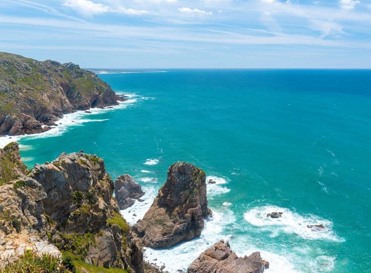Rocky cliffs meet the turquoise ocean under a blue sky with wispy clouds. Waves crash against the rocks.