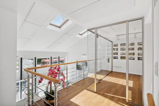 Helle Büroräume mit Glaswänden, Holzböden, Oberlichtern und gerahmten Bildern an der Wand. In der Ecke steht ein rosa Blumenarrangement.