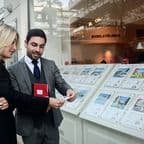 A woman and a man in business attire discuss in front of a real estate office window displaying property listings.