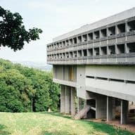 Exterior view of a modernist concrete building designed by Le Corbusier, featuring a distinctive brutalist style with repetitive window patterns and elevated structure on pillars. The building is surrounded by green landscape with trees, showcasing the integration of modern architecture with nature.