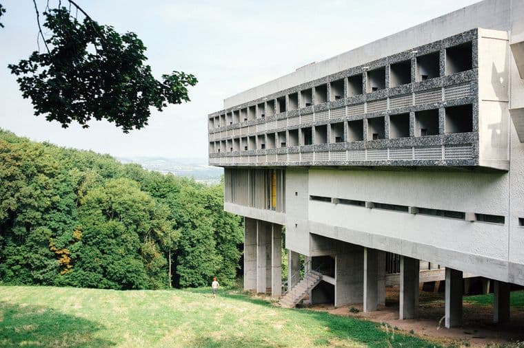 Exterior view of a modernist concrete building designed by Le Corbusier, featuring a distinctive brutalist style with repetitive window patterns and elevated structure on pillars. The building is surrounded by green landscape with trees, showcasing the integration of modern architecture with nature.
