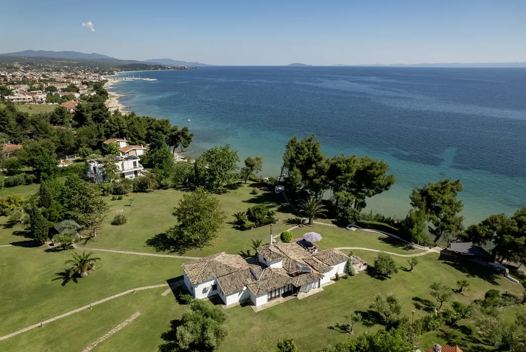 Coastal village with white houses and red-tiled roofs on a grassy hillside, surrounded by trees, overlooking turquoise bay that fades to blue at the horizon.