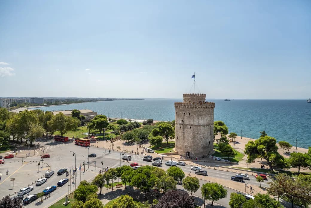 Das Stadtbild von Thessaloniki an der Küste zeigt den ikonischen Weißen Turm am Meer, umgeben von Bäumen und belebten Straßen mit Autos und Bussen. Der Himmel ist klar und sonnig, während sich das endlose Meer am Horizont erstreckt.