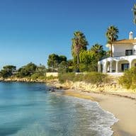 Ground-level view of a white Mediterranean-style villa with arched porticos and balconies, positioned on a rocky coastline. The property features tall palm trees and colorful bougainvillea gardens, overlooking a secluded sandy beach with gentle waves from turquoise waters.