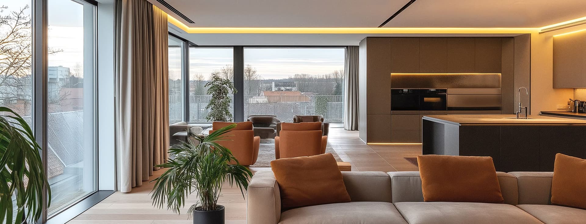 Luxurious penthouse living space featuring floor-to-ceiling windows, a grey L-shaped sofa, tan leather armchairs, and an open-plan kitchen. The room is decorated with indoor plants and illuminated by warm LED ceiling lights, showcasing a contemporary design with neutral colors and natural light.