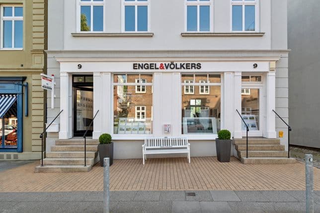 Straßenansicht des Engel & Völkers-Immobilienbüros mit einer weißen Bank und Topfpflanzen im Außenbereich, mit großen Fenstern, in denen Angebote angezeigt werden.