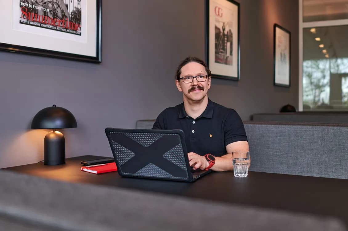 Man with glasses and a mustache sits at a desk with a laptop in a modern office lounge, framed posters on the wall.