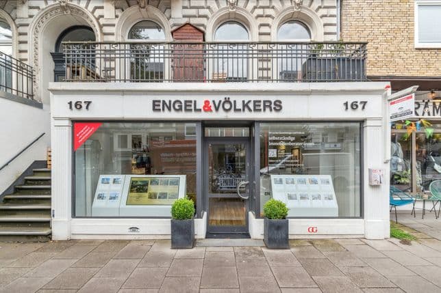 Schaufenster von Engel & Völkers mit großen Schaufenstern, die Immobilienangebote anzeigen, flankiert von zwei Topfpflanzen und einer Treppe auf der linken Seite.