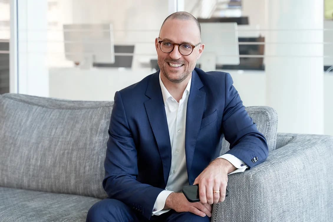 Smiling man in a navy suit and white shirt with round glasses sits on a gray patterned sofa in a bright office, holding a smartphone.