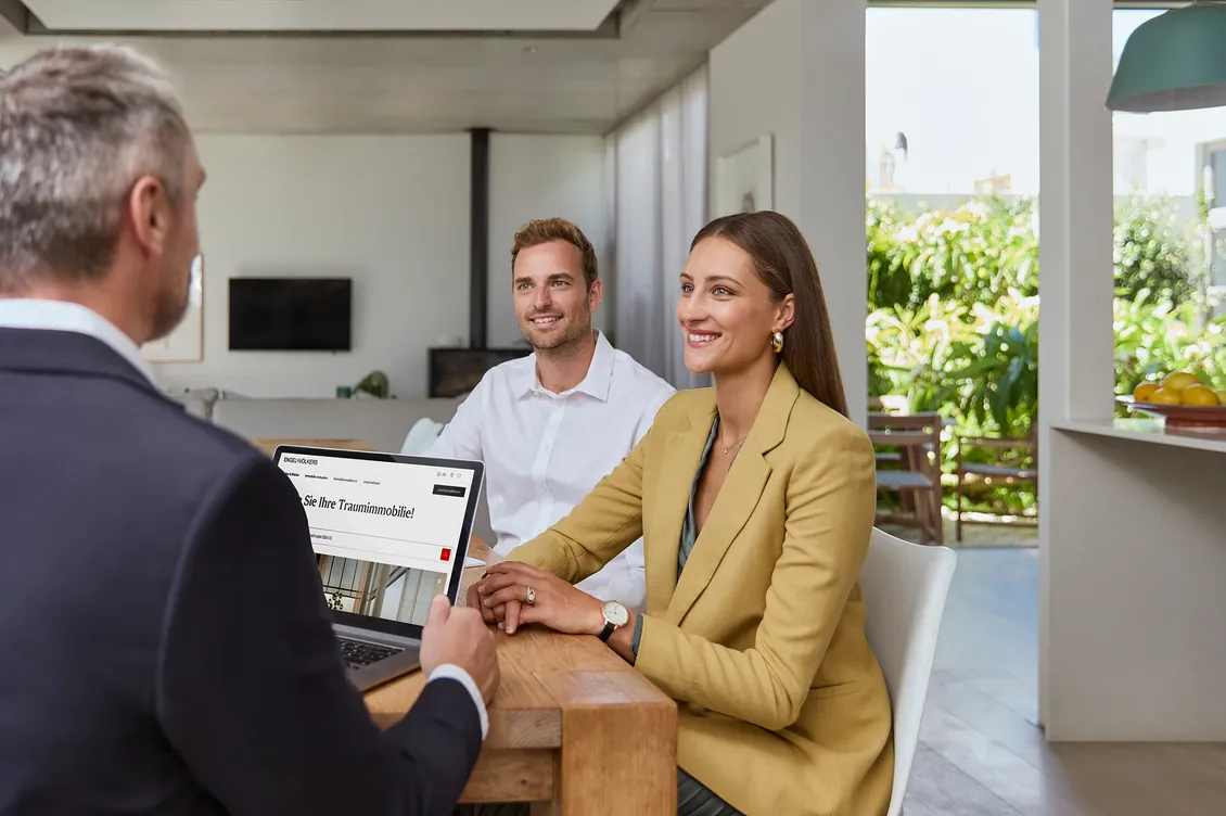 Ein Paar in einer Finanzierungsberatung bei Engel & Völkers Finance. Sie sitzen mit einem Berater an einem Tisch in einem modernen Wohnraum und blicken lächelnd auf seinen Laptop.