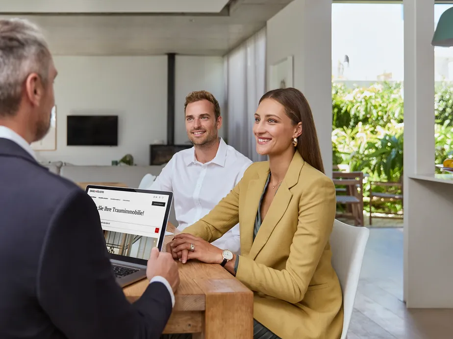 Ein Paar in einer Finanzierungsberatung bei Engel & Völkers Finance. Sie sitzen mit einem Berater an einem Tisch in einem modernen Wohnraum und blicken lächelnd auf seinen Laptop.