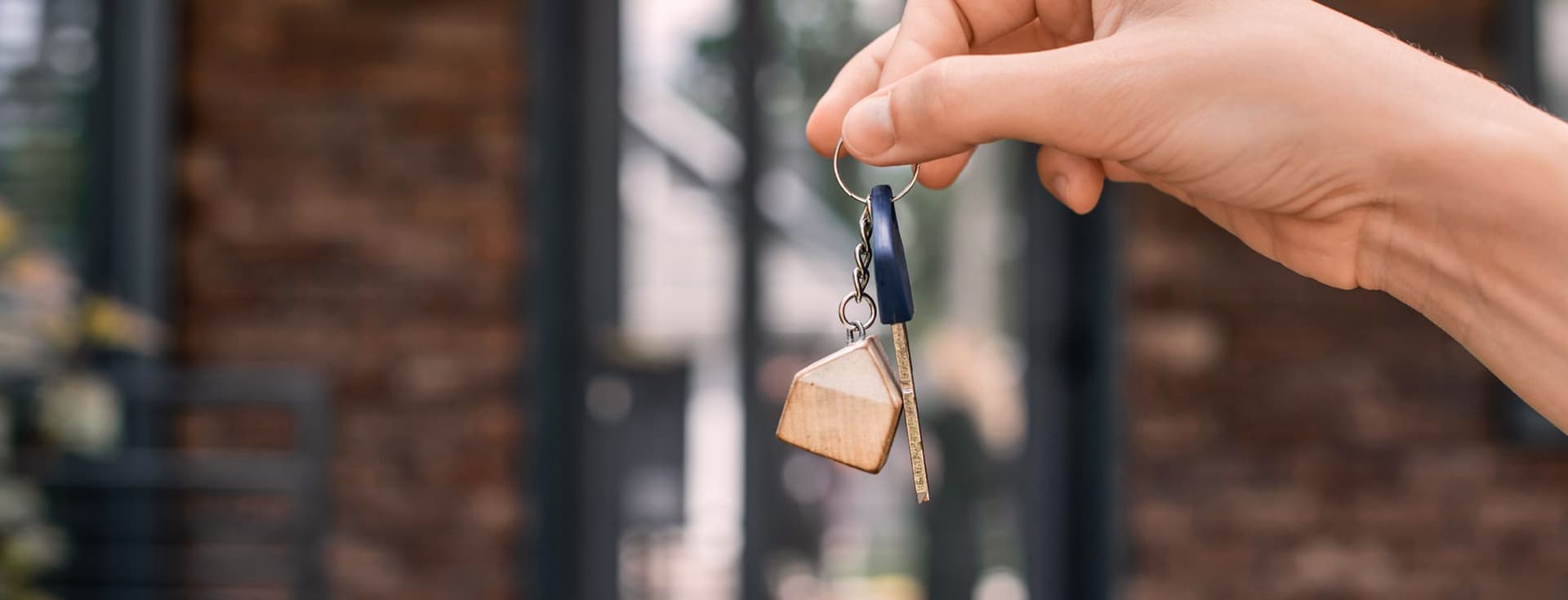 Hand holding a key with a house-shaped keychain in front of a brick building entrance.