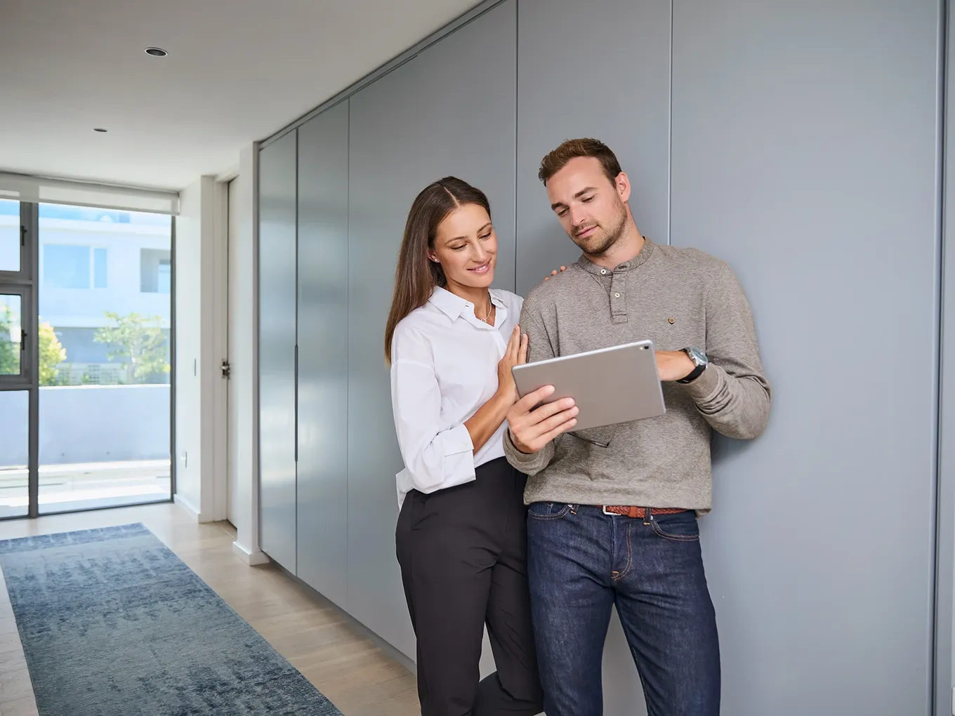 Ein Mann und eine Frau stehen in einer hellen, modernen Wohnung und lächeln, während sie auf den Bildschirm eines Tablets schauen, das der Mann in den Händen hält.