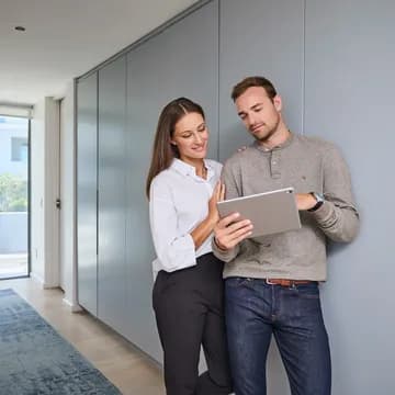A man and a woman stand in a bright, modern apartment, smiling as they look at the screen of a tablet held by the man.