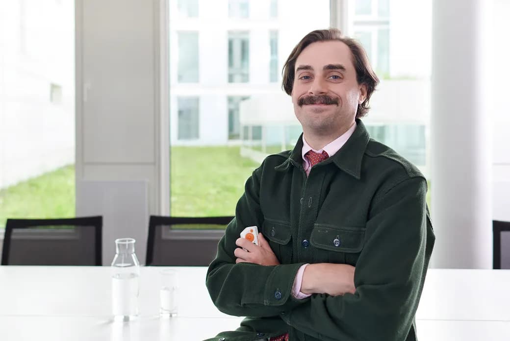 Smiling man with a mustache in a green jacket sits at a bright conference table, arms crossed, holding a small orange device; large windows behind.