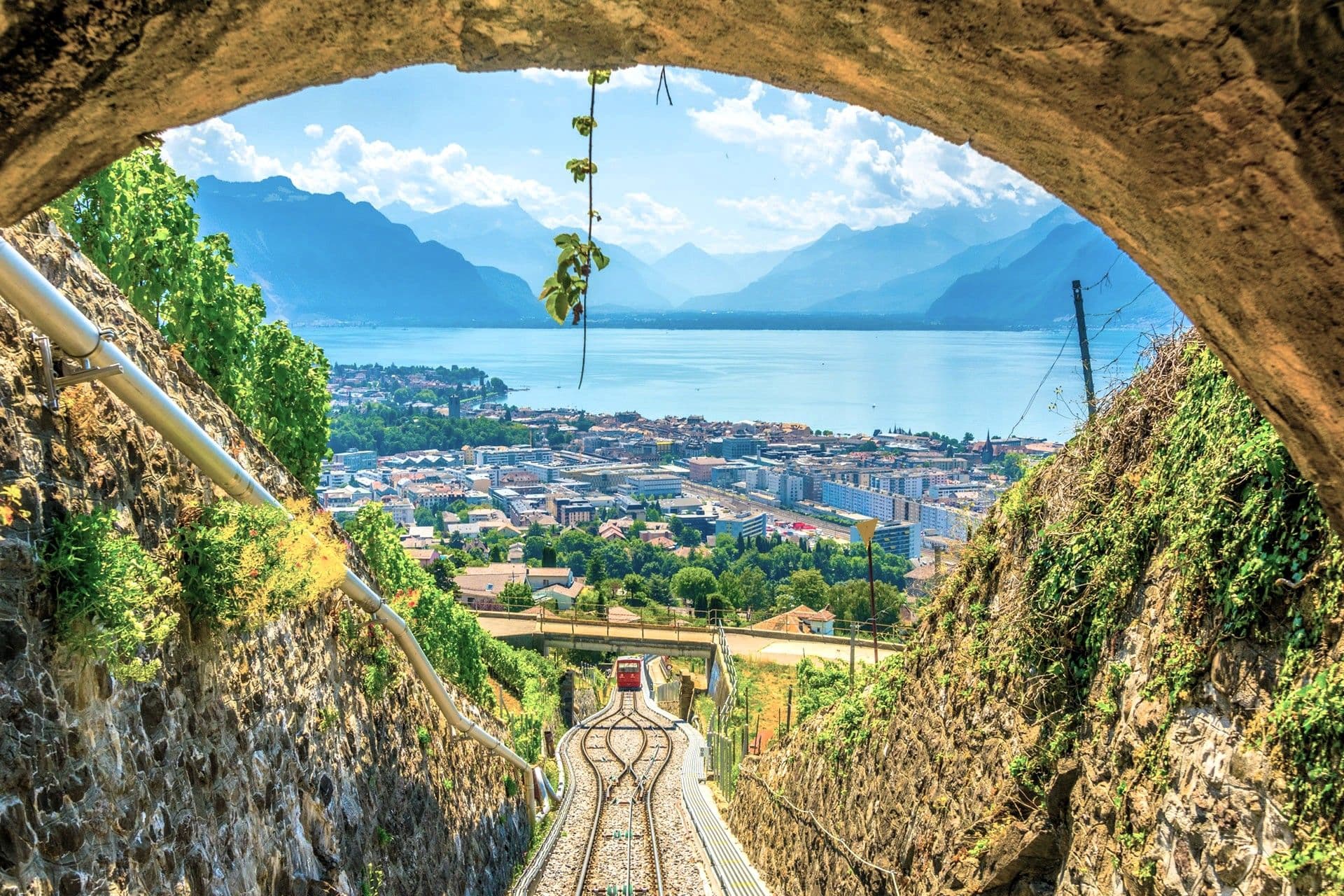 Rail tracks descend through a stone tunnel, opening to a lakeside town with blue mountains in the distance under a bright sky.