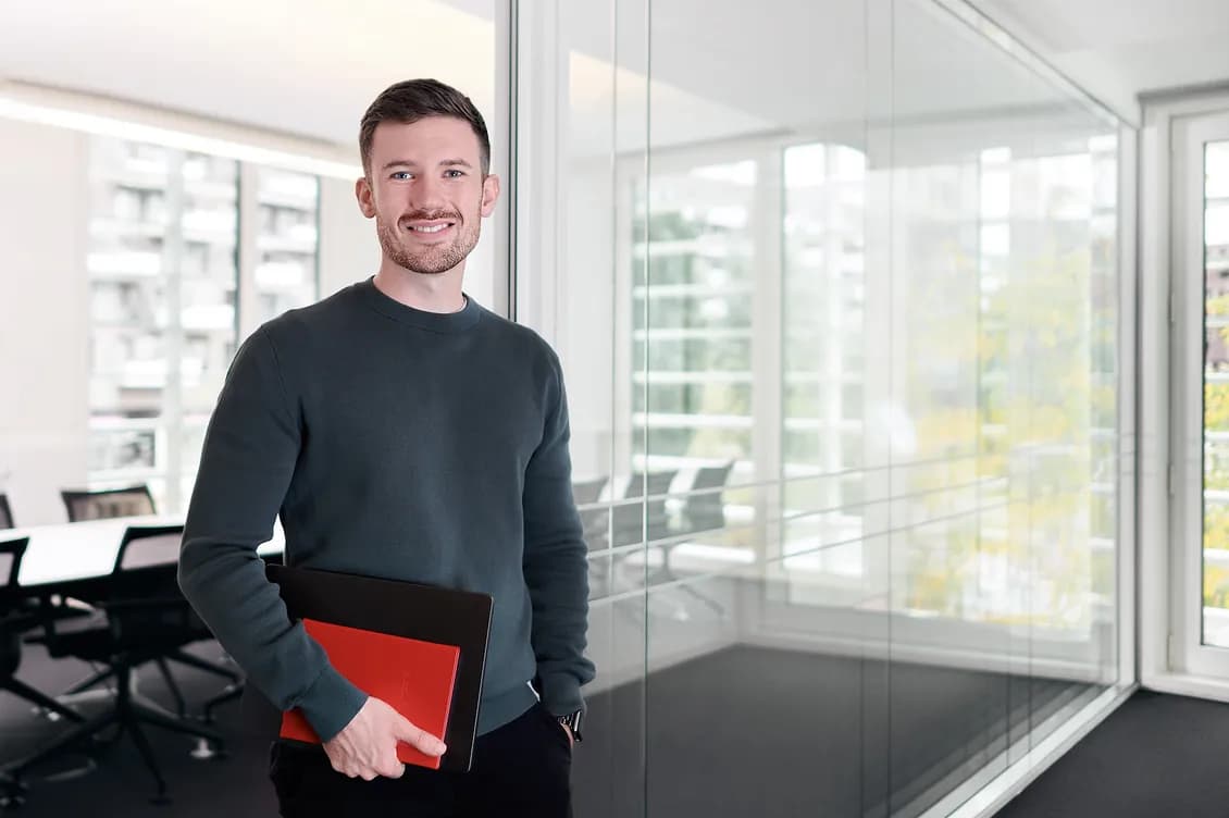 Smiling man in a dark sweater stands beside glass walls in a bright office, holding a red notebook and black folder.