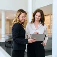 Zwei Frauen im Business-Outfit in einem Büro schauen gemeinsam auf ein Tablet.
